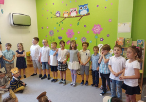 W sali przedszkolnej w półkolu stoi grupa elegancko ubranych dzieci. Niektóre z nich mają otwarte usta. W tle widać zieloną ścianę z kolorowymi naklejkami z czterema sowami.